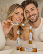 casal sorrindo com rostos juntos mulher mostrando frasco de perfume portátil feito de vidro âmbar com tampa prateada na horizontal com mão direita e três frascos sob pedra de granito clara