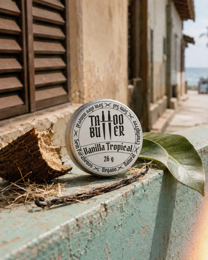 Latinha de alumínio Matchii Vanilla Tropical com rótulo branco redondo, apoiada sobre superfície desgastada, cercada por coco aberto, folha verde e fava de baunilha em cenário externo urbano.