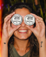 Mulher sorridente segura duas latinhas de alumínio Matchii diante dos olhos, com rótulos brancos redondos escritos Cupuaçu Cream e Neroli Sunny, em ambiente externo com vegetação ao fundo.