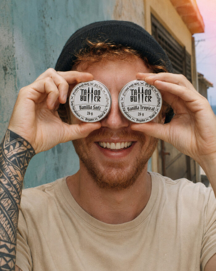Homem sorridente segura duas latinhas de alumínio Matchii diante dos olhos, com rótulos brancos redondos Vanilla Soft e Vanilla Tropical, em cena externa com rua ao fundo.