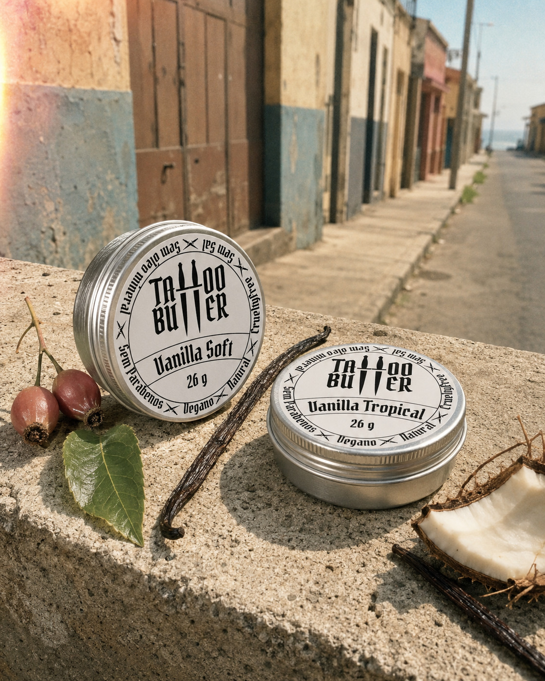 manteiga-para-tatuagem-tattoo-butter-vanilla-soft-vanilla-tropical Duas latinhas de alumínio Matchii, com rótulos brancos redondos escritos Vanilla Soft e Vanilla Tropical, apoiadas sobre muro de concreto com baunilha, coco, folha e frutos ao redor.